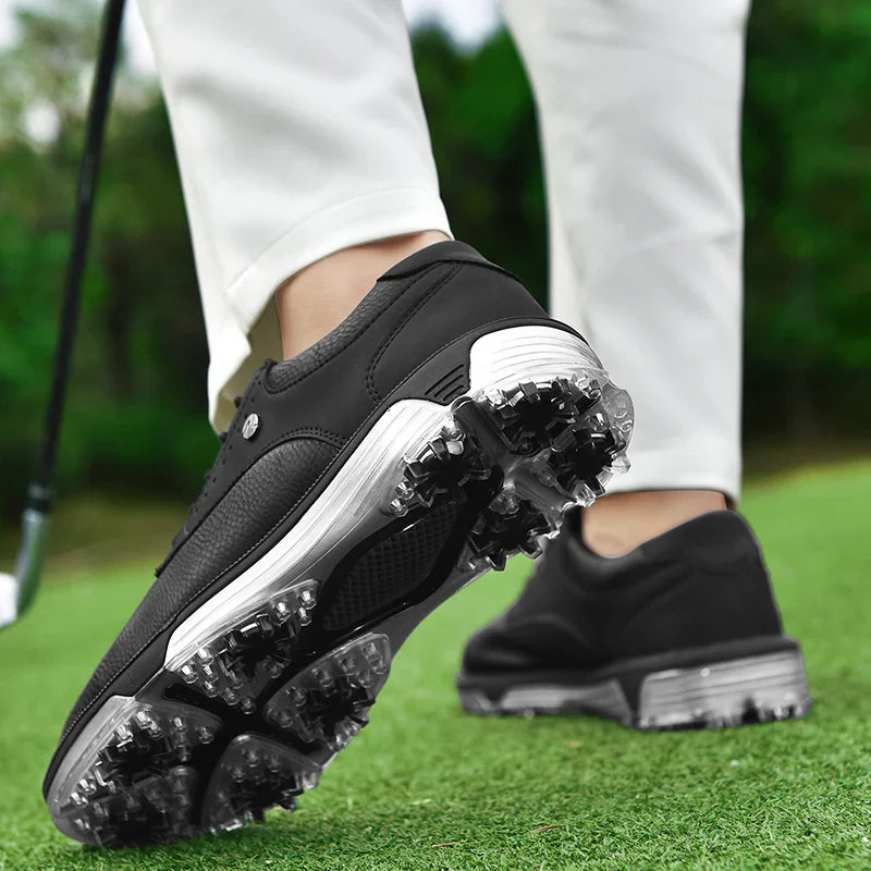 Close-up of a person wearing black waterproof golf shoes with spikes, standing on green grass. The person is holding a golf club, and only their legs and shoes are visible. Trees are blurred in the background.