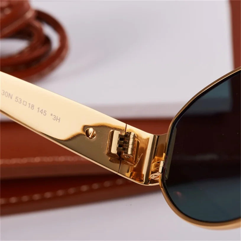 Close-up of vintage-inspired sunglasses with a gold frame, highlighting the hinge and arm details engraved with specifications. A blurred brown leather case and white surface set an elegant Italian style backdrop.