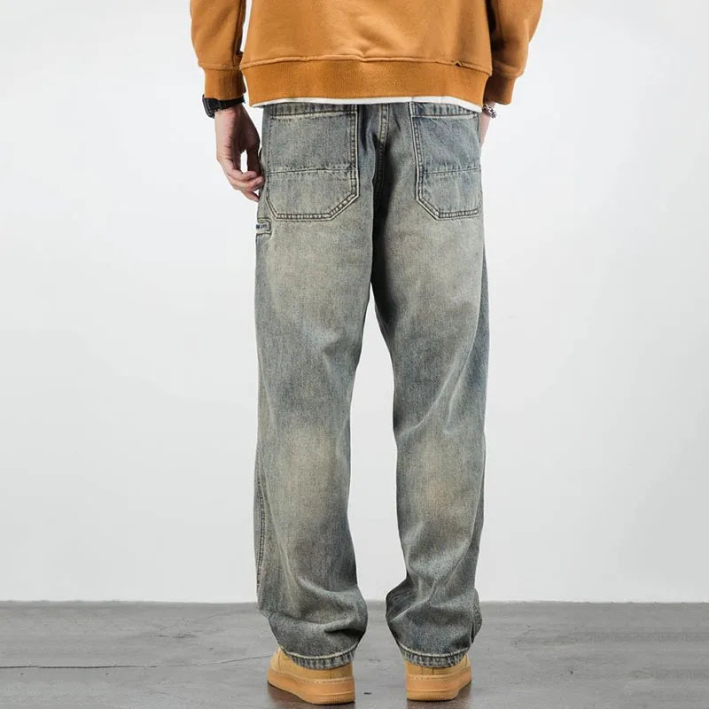 A person stands facing away, wearing a brown sweatshirt, loose-fitting stone washed jeans, and tan shoes, against a plain light background.