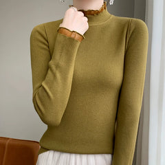 A person wearing an olive green ribbed cashmere blend turtleneck sweater with sheer ruffle details at the neck and cuffs, paired with a light pleated skirt, stands indoors near a brown chair.
