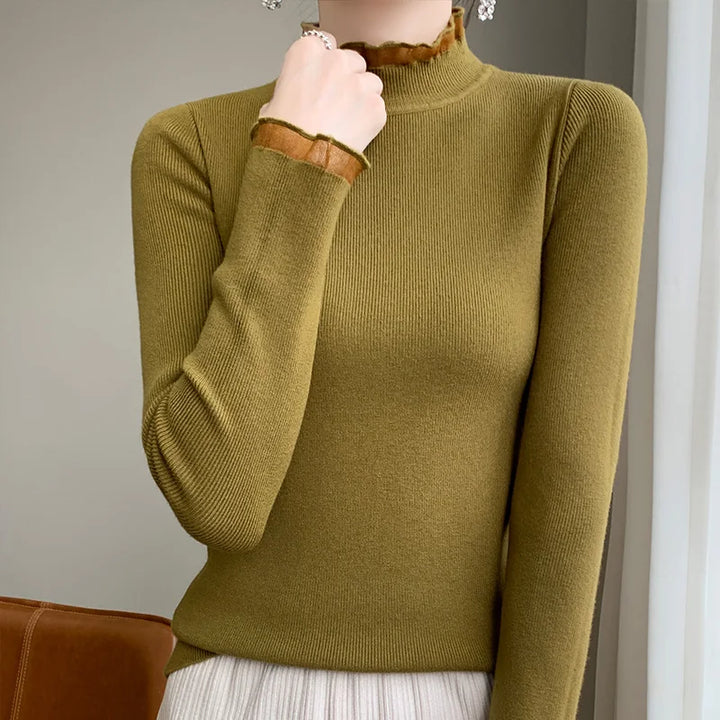 A person wearing an olive green ribbed cashmere blend turtleneck sweater with sheer ruffle details at the neck and cuffs, paired with a light pleated skirt, stands indoors near a brown chair.