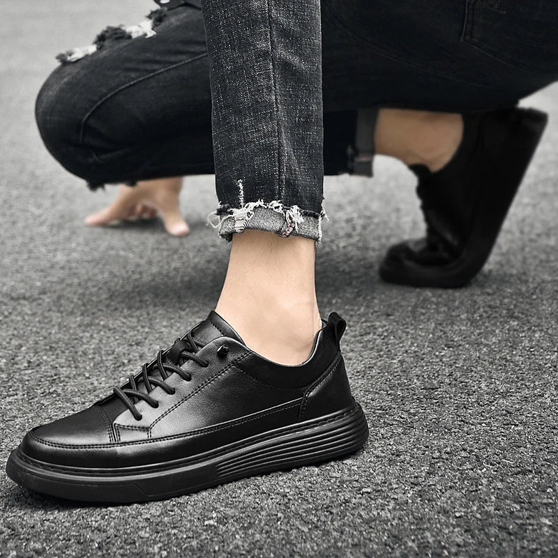 A person wearing black leather sneakers with genuine leather and distressed, cuffed jeans is crouching on a gray asphalt surface. The focus is on the stylish shoes and rough texture of the jeans’ hem.