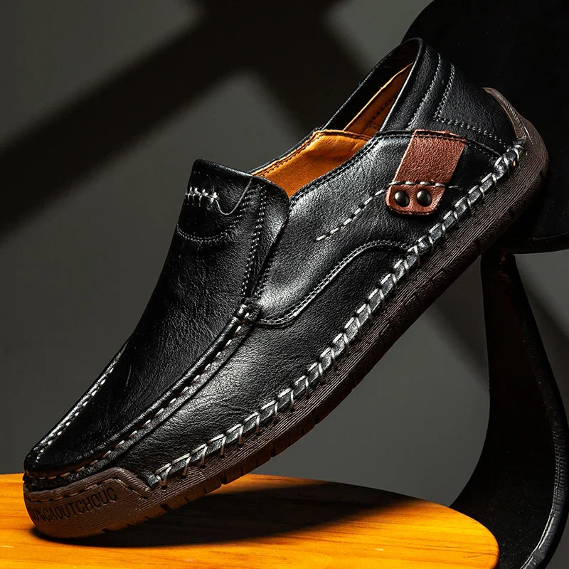 A black leather loafer with detailed stitching, a decorative brown accent on the side, and a textured rubber sole, displayed at an angle on a wooden surface against a dark background.