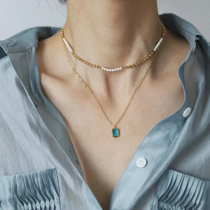 A person wearing a light blue, silky blouse and three layered necklaces: a pearl choker, an 18K gold plated chain, and a longer gold chain with a rectangular green zirconia pendant. The blouse features button details and a pocket flap.
