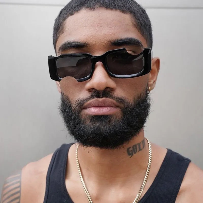 A man with a short beard and mustache wearing geometric frames sunglasses, a black tank top, and a gold chain necklace. He has a tattoo on his neck, embracing street style as he stands against a plain background.