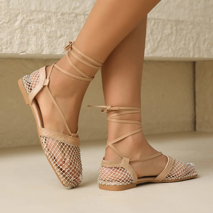 A person wearing beige flat sandals with a mesh upper and ankle-wrap straps, standing against a light stone background. The sandals feature closed toes, open sides, and a slip-resistant sole for added comfort and stability.