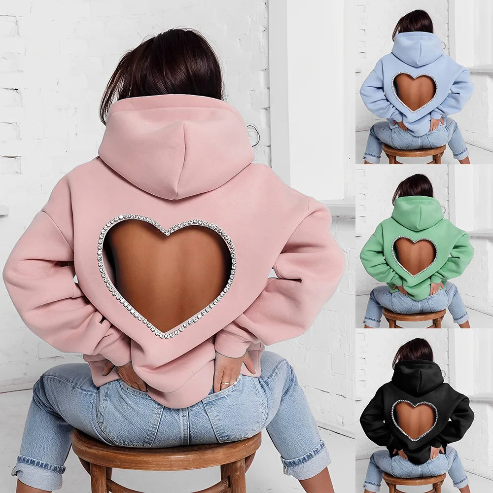 A person wears an oversized, heart-shaped open back hoodie with a large cutout revealing skin. Available in pink, blue, green, and black. The person sits on a stool with their back facing the camera. Features optional rhinestone trim for extra sparkle.