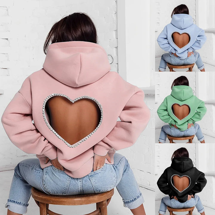 A person wears an oversized, heart-shaped open back hoodie with a large cutout revealing skin. Available in pink, blue, green, and black. The person sits on a stool with their back facing the camera. Features optional rhinestone trim for extra sparkle.