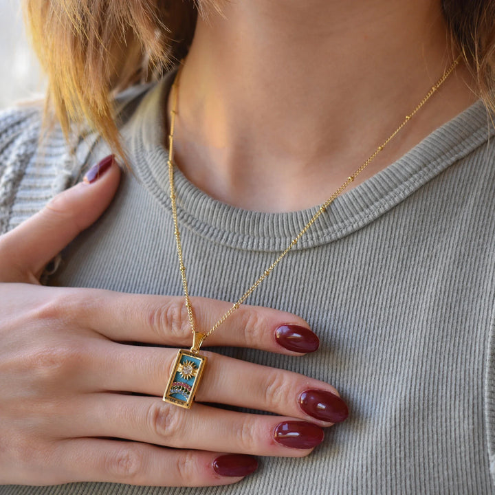 A woman with maroon painted nails wears the Solaris Pendant Necklace, an 18K gold-plated piece with blue enamel and gold details, resting on a ribbed light gray top. One hand is gently touching her chest near the pendant.