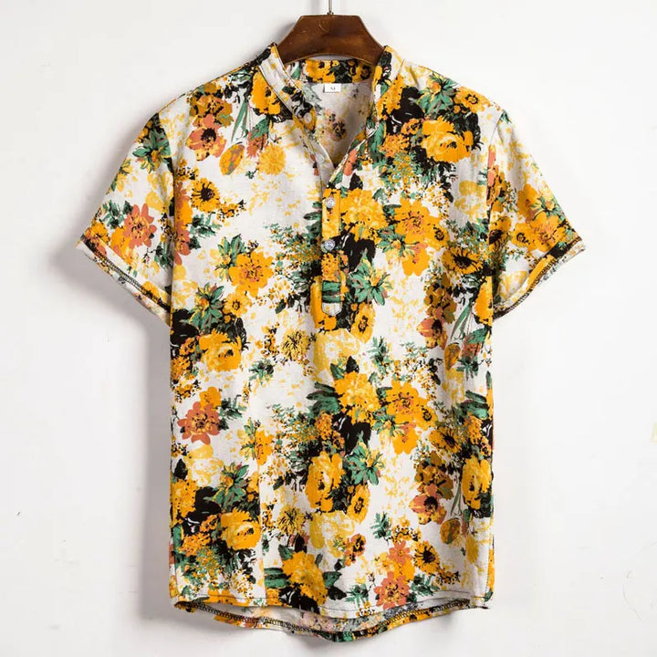 A short-sleeved cotton shirt with a vibrant floral pattern in yellow, orange, green, and black on a white background, hanging on a brown wooden hanger against a plain white wall.