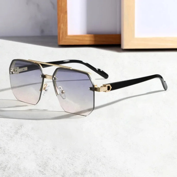 A pair of geometric, oversized sunglasses with a gold geometric metal frame and gradient UV400 protective lenses rests on a light surface, with a blurred wooden picture frame in the background.