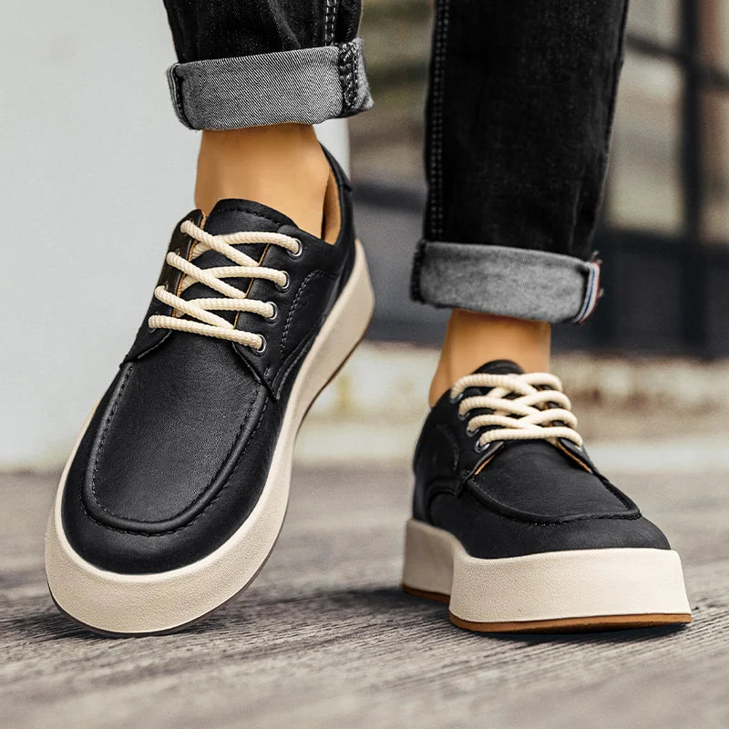 A person wearing black lace-up sneakers with thick white soles and cream-colored laces, reminiscent of Italian designer shoes, stands on a wooden surface. The person's jeans are rolled up at the ankles.