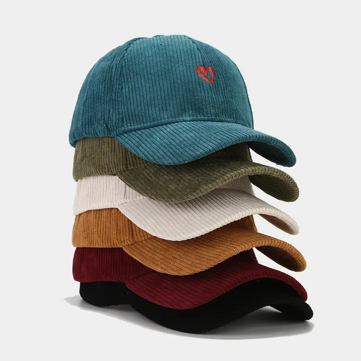 A stack of six corduroy hats in teal, olive green, beige, orange-brown, burgundy, and black. The top cap features a small red heart embroidery on the front for a charming touch.