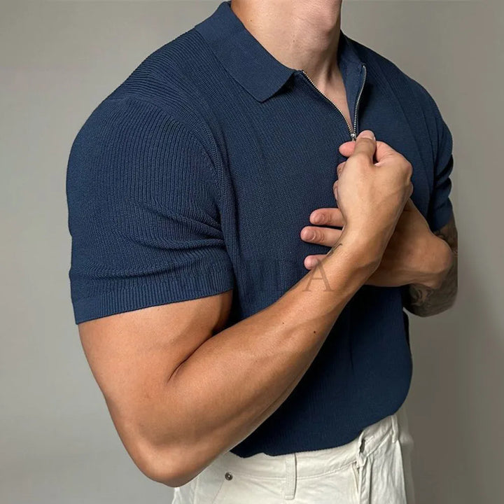 A person wearing a navy blue quarter zip polo with ribbed detailing and off-white pants stands against a plain background, with one hand gripping the zipper near their chest.