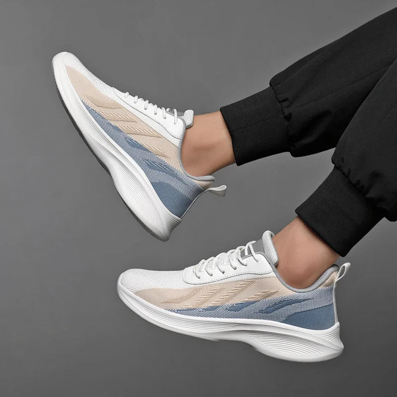 A person wearing black pants and stylish workout shoes with arch support, featuring white soles and beige, blue, and gray accents, against a plain gray background. The feet are lifted mid-air, highlighting the shoes’ sleek design.