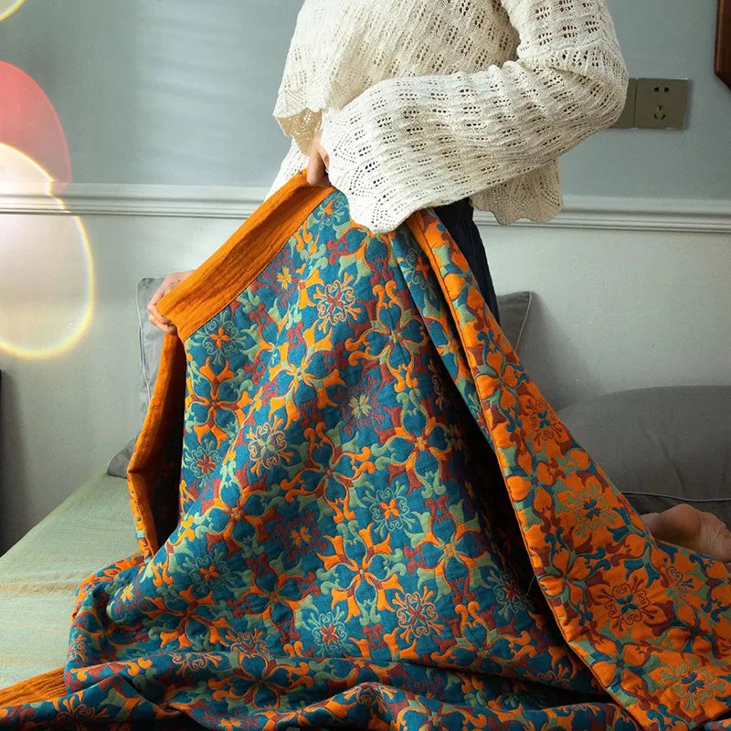 A person in a cream knit sweater holds a vibrant ethnic pattern blanket with orange, blue, and green floral designs while sitting on a couch. Sunlight creates a soft lens flare effect in the background.