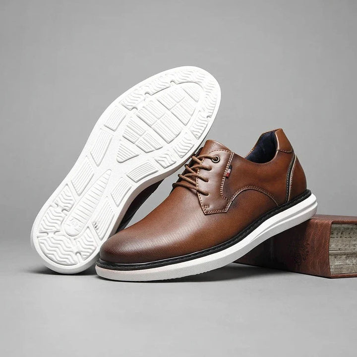 A pair of brown genuine leather oxford lace-up shoes with traditional detailing and white rubber soles, one upright and the other on its side to display the textured sole pattern, set against a gray background.