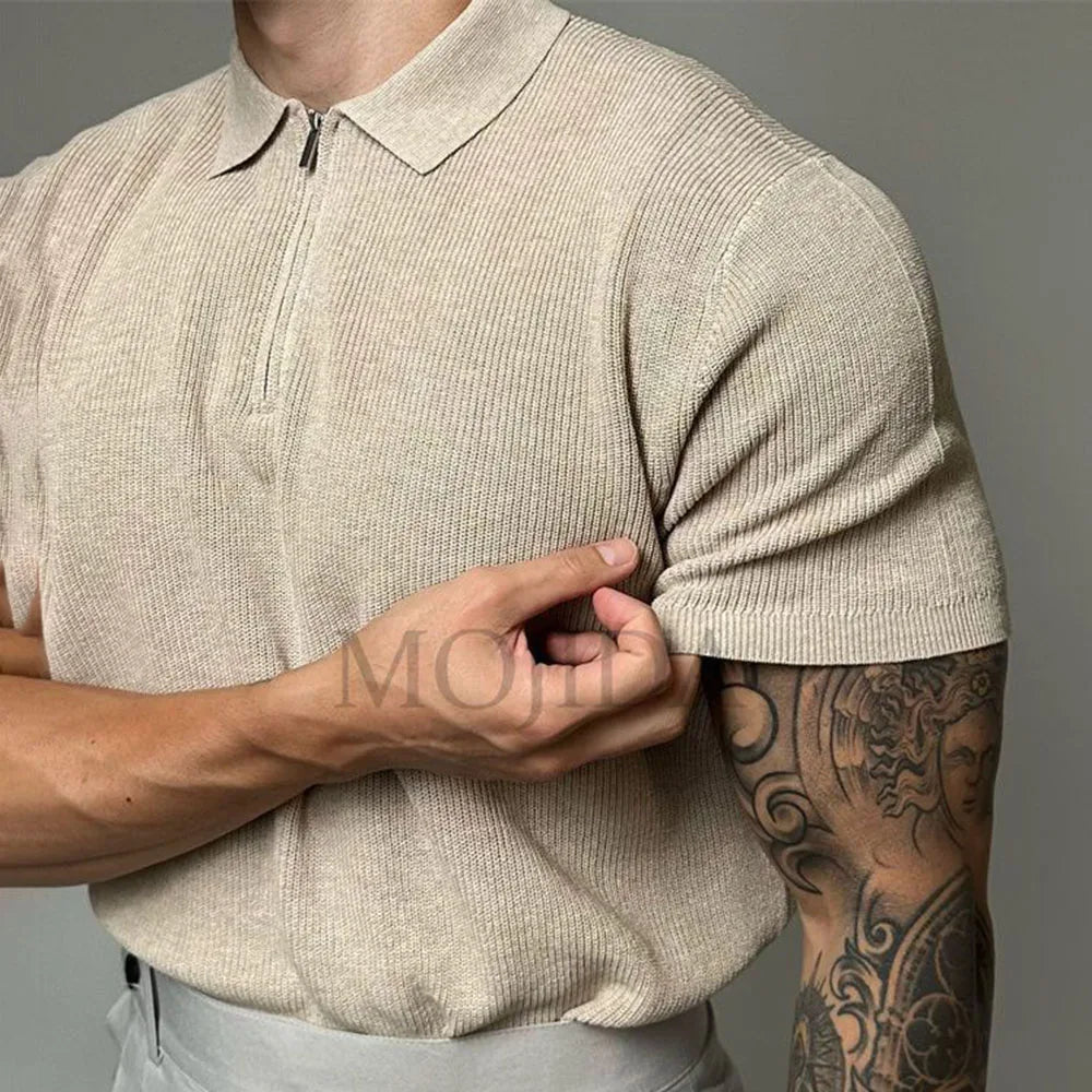 A person wearing a beige viscose polo with ribbed detailing and a zipper collar is pulling up their sleeve, revealing a detailed tattoo on their arm. The background is plain and neutral.