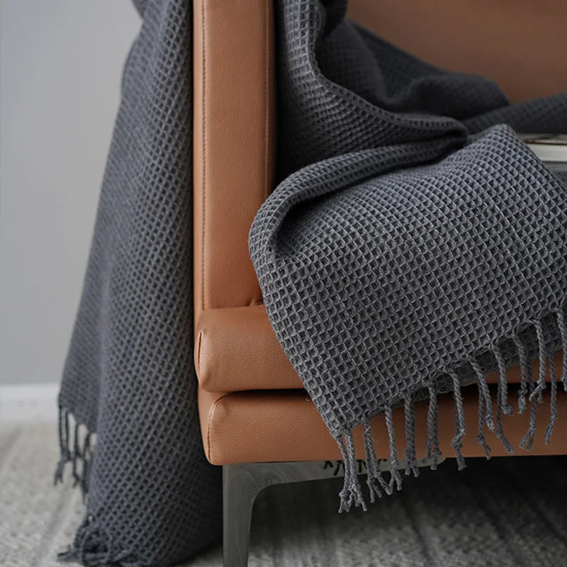 A close-up of a brown leather chair draped with a dark gray knit waffle blanket with fringe, set on a light gray patterned rug—a perfect decorative accent blanket for cozy spaces.