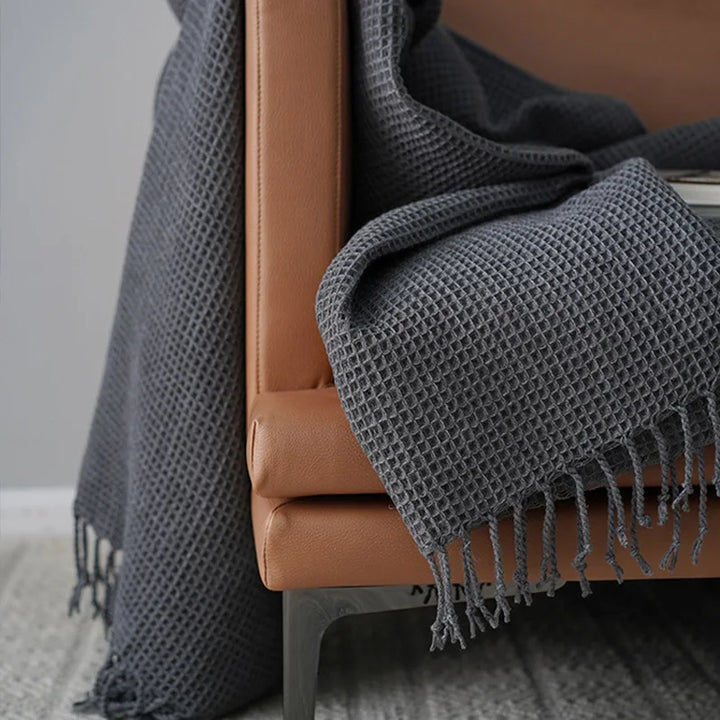A close-up of a brown leather chair draped with a dark gray knit waffle blanket with fringe, set on a light gray patterned rug—a perfect decorative accent blanket for cozy spaces.
