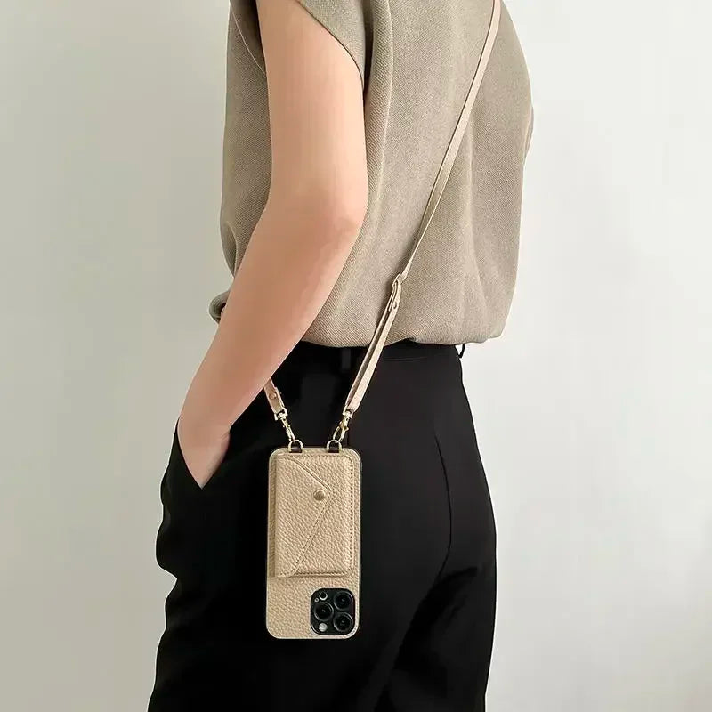 A person wearing a beige top and black pants carries a beige crossbody phone case crafted from pebble-texture material with a small pouch attached. The phone case hangs by their side against a plain light background.
