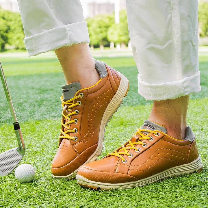 A person wearing brown golf footwear with yellow laces, featuring advanced cleat technology, stands on green grass next to a golf ball and club. Their white pants are rolled up, with trees and buildings blurred in the background.