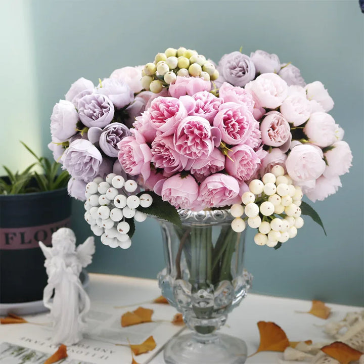 A glass vase holds a bouquet of pink and light purple peonies with faux petals, accented by white berries, set on a table with scattered dried leaves, a small angel figurine, and a potted plant in the background—perfect for maintenance-free decor.