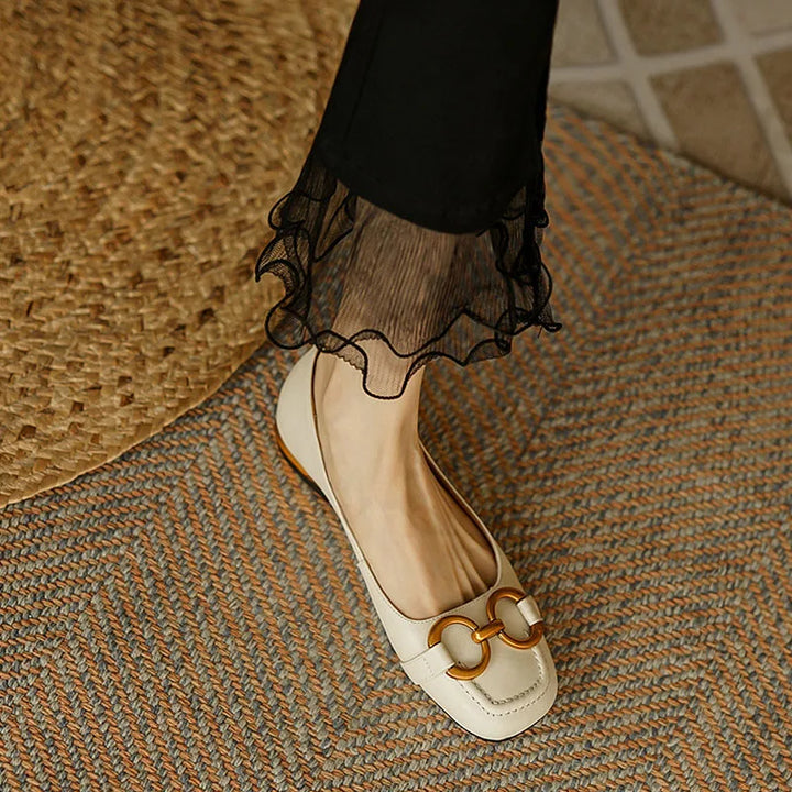 A person wearing black pants with sheer ruffled hems and cream-colored loafers featuring a gold buckle and cushioned insole, standing on a textured woven rug.
