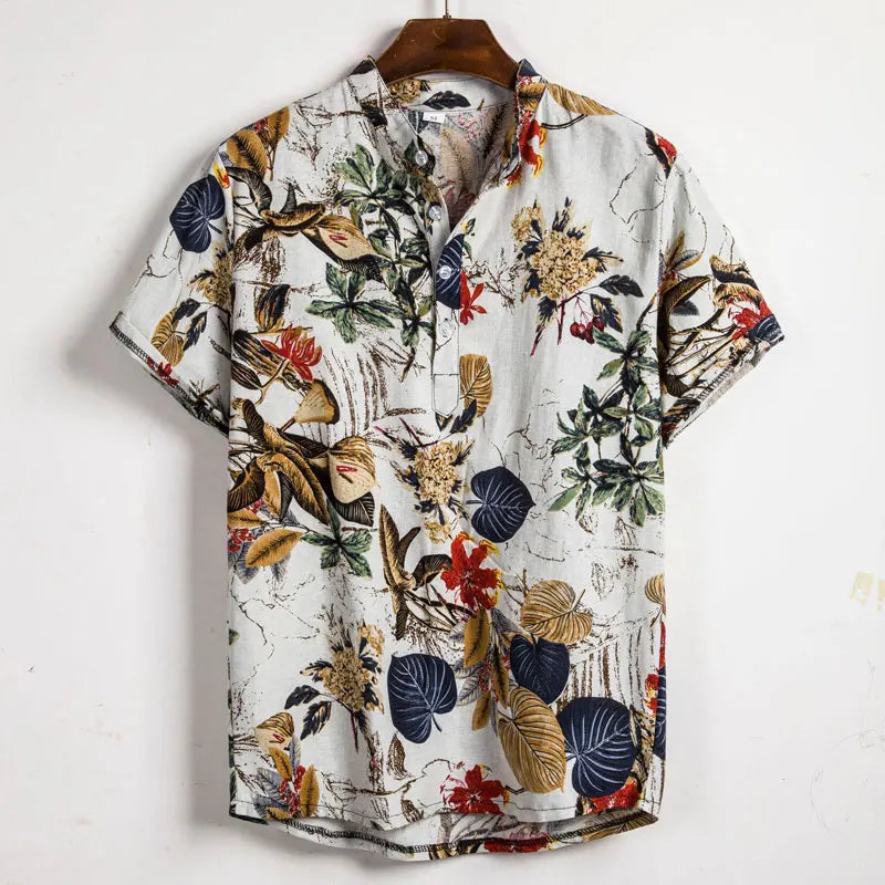 Short-sleeve button-up cotton shirt with a white background and a colorful tropical floral pattern, featuring leaves and flowers in green, navy, yellow, and red—a classic design hanging on a wooden hanger.