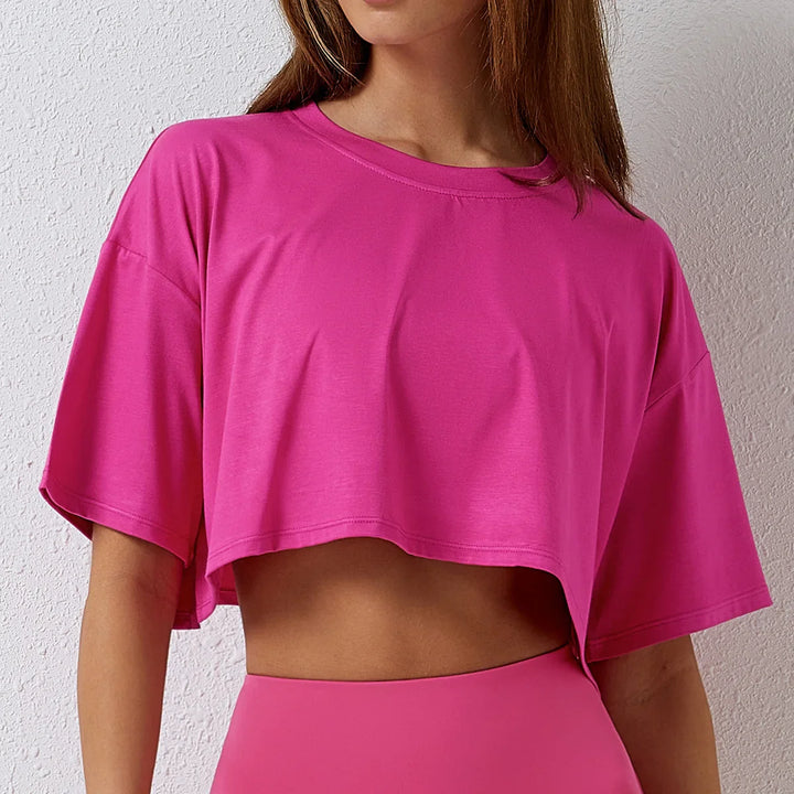 A woman in a bright pink cotton crop top with a relaxed fit and matching high-rise pants stands against a textured white wall. Her face is partially out of frame.