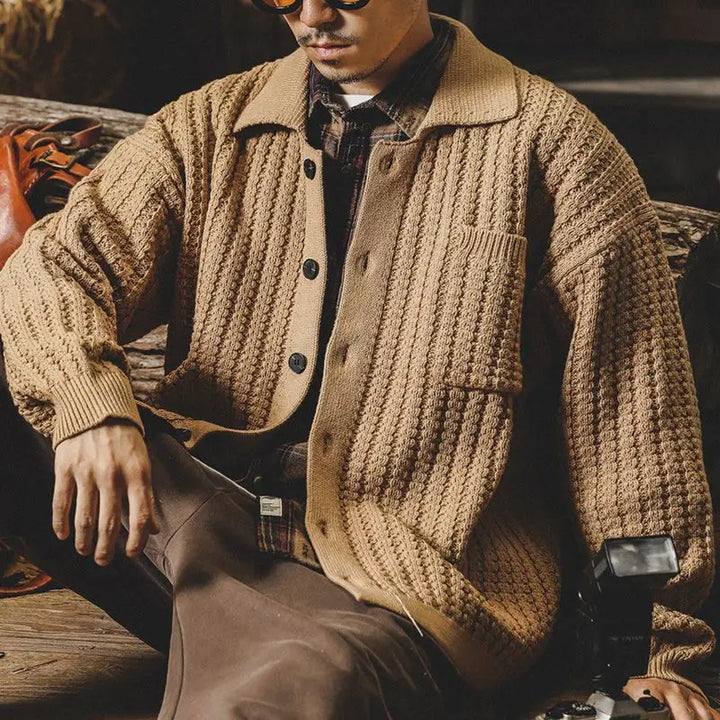 Man sitting on a wooden bench, wearing a chunky knit beige cardigan over a plaid shirt and brown pants, with sunglasses and a camera resting by his side, channeling a relaxed heritage aesthetic.