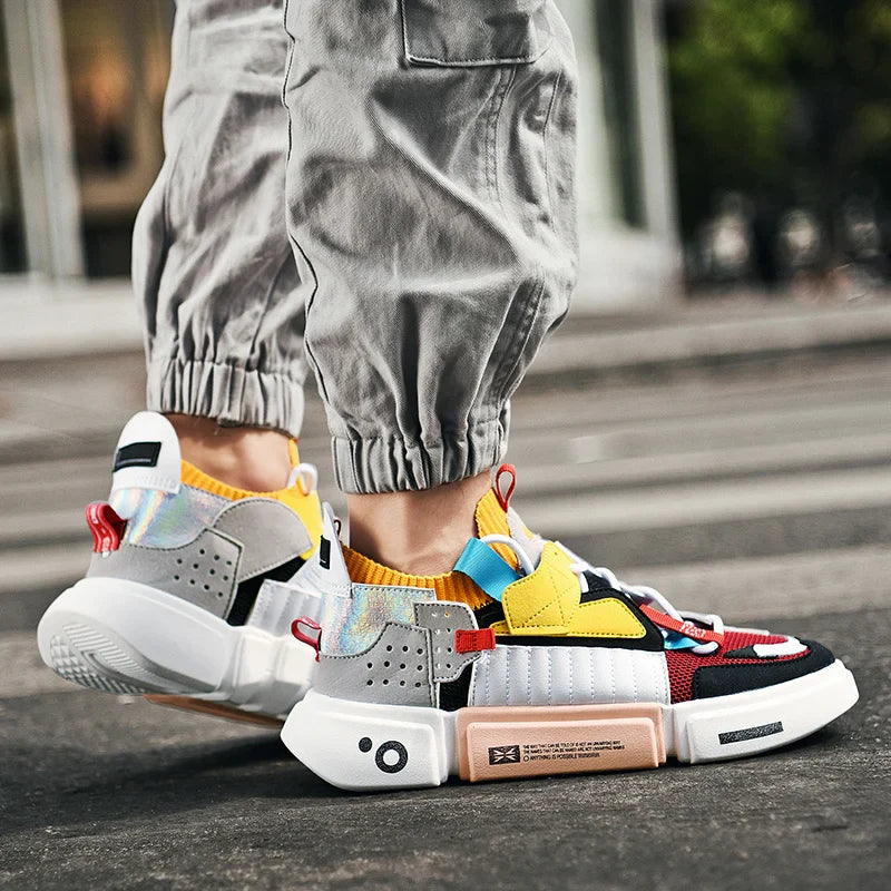 A person wearing colorful Extreme VS7 Sneakers with yellow, red, blue, gray, and white accents stands on a city street. Their lightweight breathable streetwear sneakers pair perfectly with light gray jogger pants slightly rolled at the ankles.