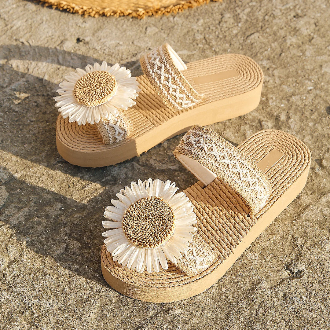Beige platform sandals with woven texture, adorned with large white faux daisy flowers on the top straps, make perfect summer footwear. Displayed on a textured stone surface in sunlight, they’re a chic pick for women's fashion sandals.