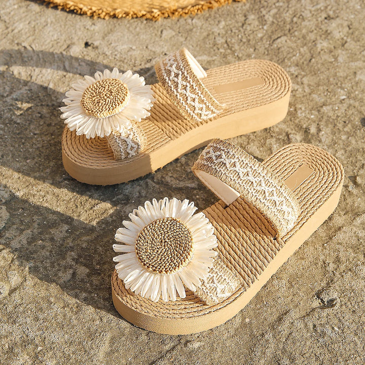 Beige platform sandals with woven texture, adorned with large white faux daisy flowers on the top straps, make perfect summer footwear. Displayed on a textured stone surface in sunlight, they’re a chic pick for women's fashion sandals.