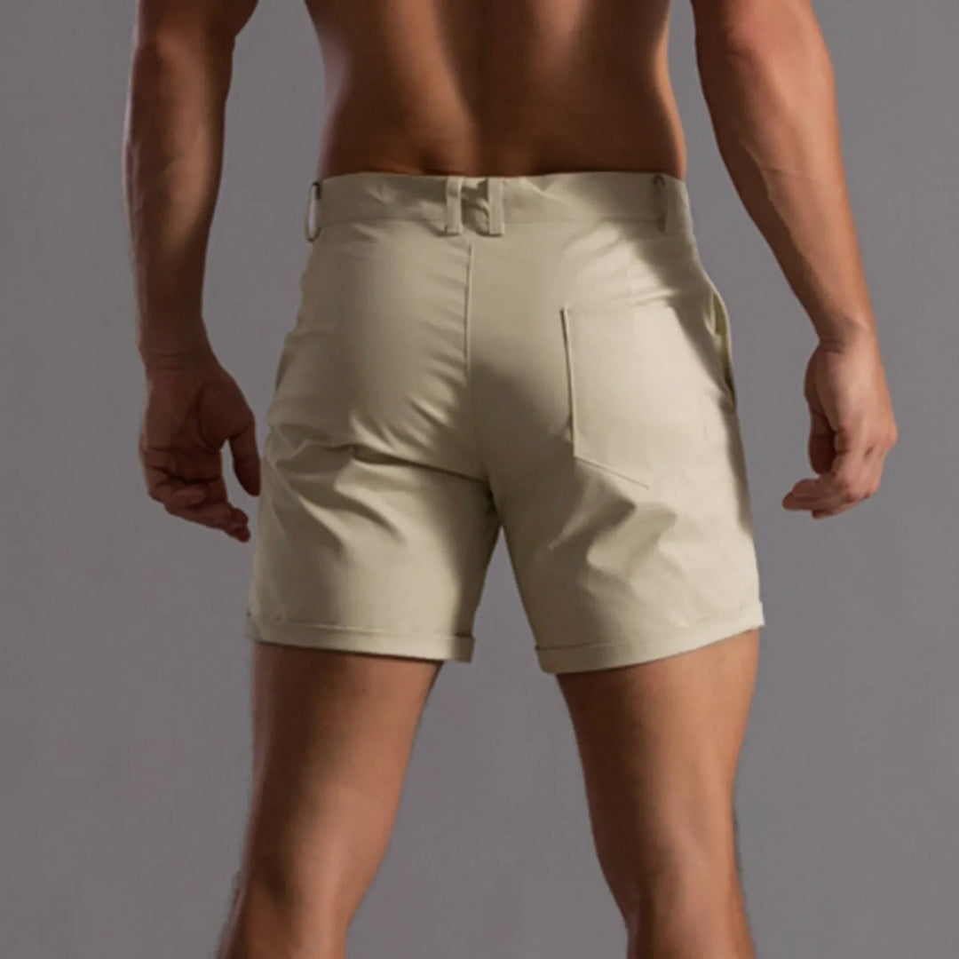 A man shown from behind wears light beige, tailored fit shorts with belt loops and rear pockets, featuring 4 way stretch, against a plain gray background.