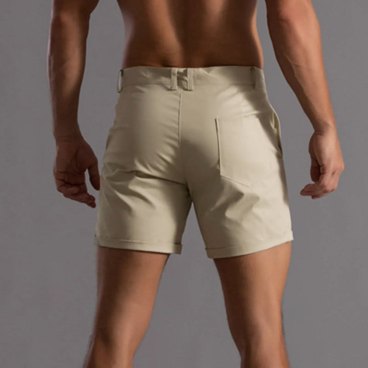 A man shown from behind wears light beige, tailored fit shorts with belt loops and rear pockets, featuring 4 way stretch, against a plain gray background.