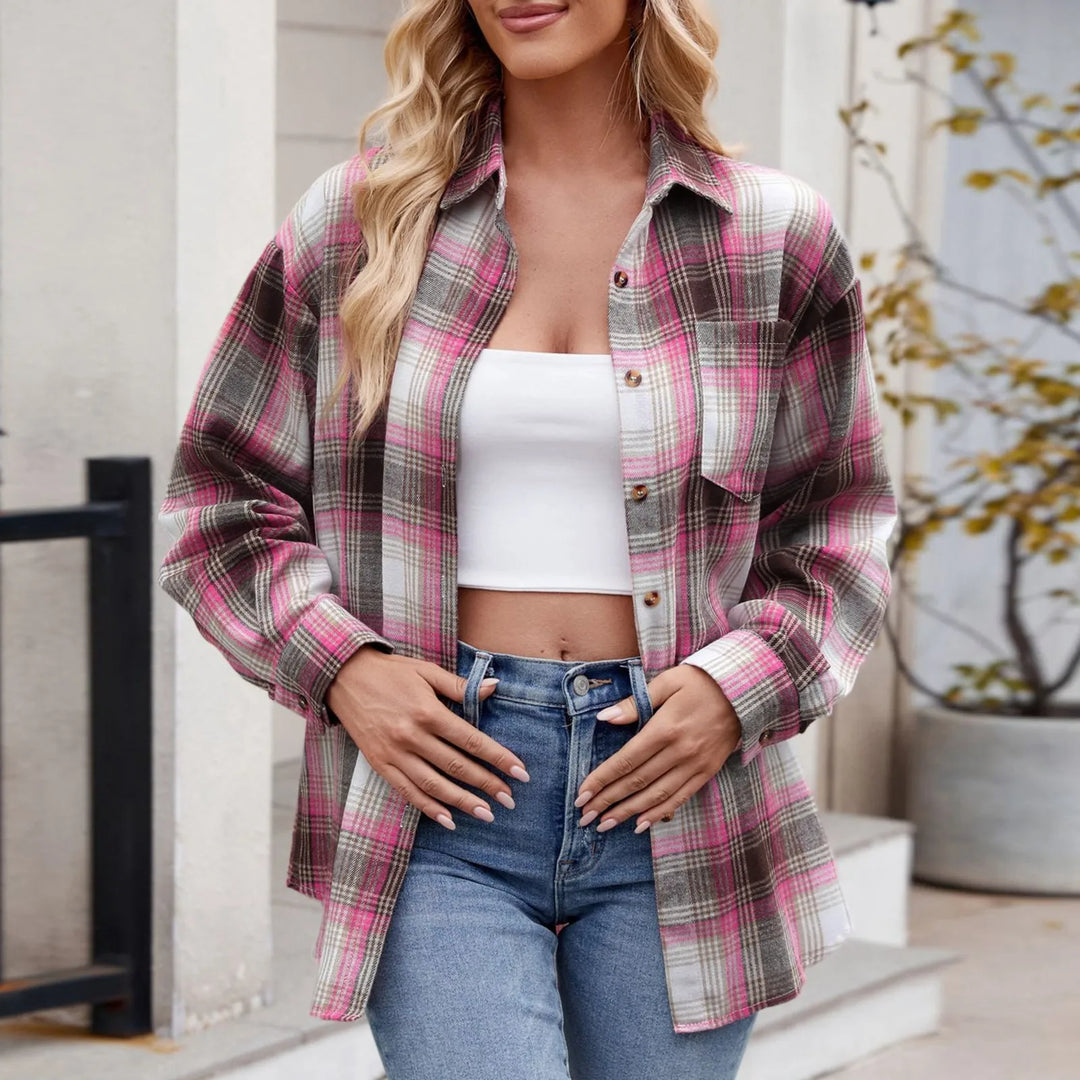 A woman stands outdoors wearing an oversized fit, pink and gray plaid button-down shirt layered over a white crop top and blue jeans. She has long, wavy blonde hair and is posing with her hands near her waist.
