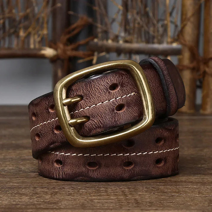 A genuine leather belt showcasing heritage craftsmanship, with white stitching and a large brass buckle, is coiled on a wooden surface against a rustic background.
