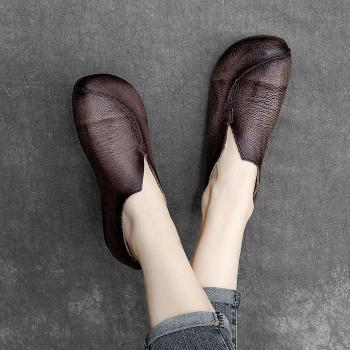 A person wearing dark brown slip-on shoes crafted from supple leather with a unique, asymmetrical design and gray jeans, legs crossed on a textured gray surface.