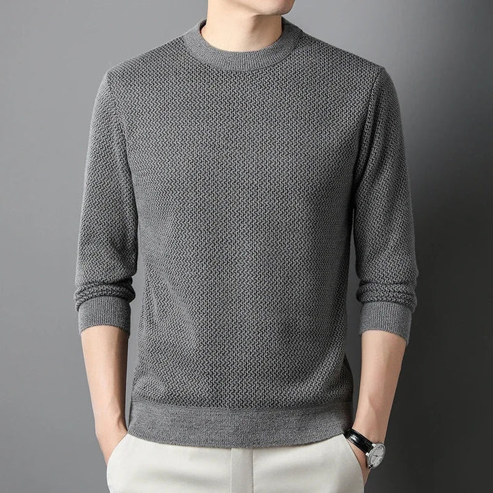 A person wearing a versatile gray knit sweater with long sleeves, light-colored pants, and a wristwatch, standing against a plain gray background. The persons head is not visible.