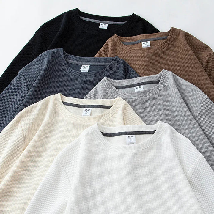 Six crewneck, waffle knit, heavyweight long-sleeve shirts in black, brown, navy, gray, cream, and white are neatly arranged on a white background. Each premium cotton shirt displays a visible white tag at the collar.