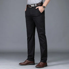 A person wearing black dress pants with a tailored fit, a light grey shirt, a brown belt, brown dress shoes, and a wristwatch, standing with hands in pockets against a plain grey background.
