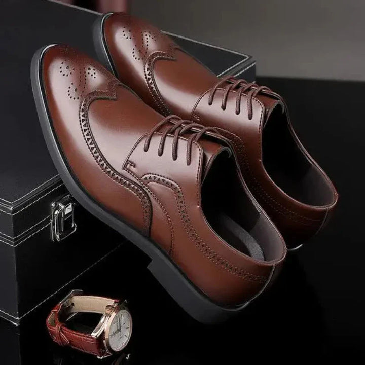 A pair of premium leather shoes with brogue detailing, featuring decorative stitching and perforations, displayed on a black surface next to a black box and a brown wristwatch with a white face.