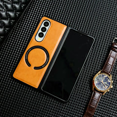 A foldable smartphone with a tan Samsung Galaxy Z Fold case lies partially open on a textured black surface, next to a brown leather strap watch and a quilted black item.