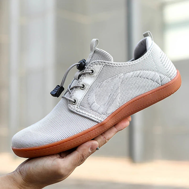A hand holds a light gray mesh sneaker with a brown rubber sole and an elastic lace tightened by a black toggle, featuring a wide toe box, displayed against a blurred outdoor background.