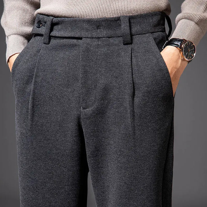 A person wearing high-waisted, dark gray tailored slacks with hands in pockets, paired with a light gray sweater and a black wristwatch. The background is dark and plain.