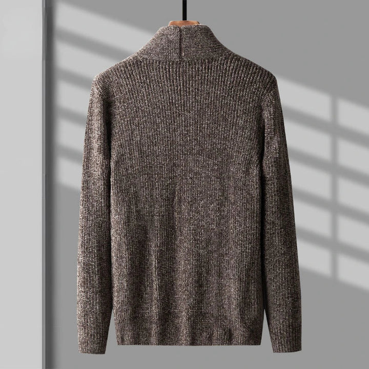 A brown, long-sleeve, ribbed knit sweater in a soft cashmere wool blend hangs on a wooden hanger against a gray wall with shadows from window blinds.