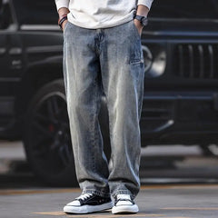 A person wearing loose-fitting, stone washed gray jeans, a white long-sleeve shirt, black wristwatch, and black-and-white sneakers stands in front of a black vehicle with hands in pockets.