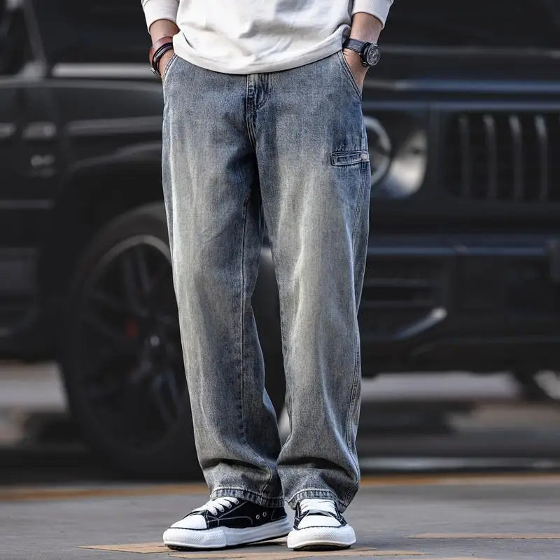 A person wearing classic fit gray denim jeans, a white long-sleeve shirt, black and white sneakers, and a watch stands outdoors in front of a black vehicle with hands in their pockets. The persons face is not visible.