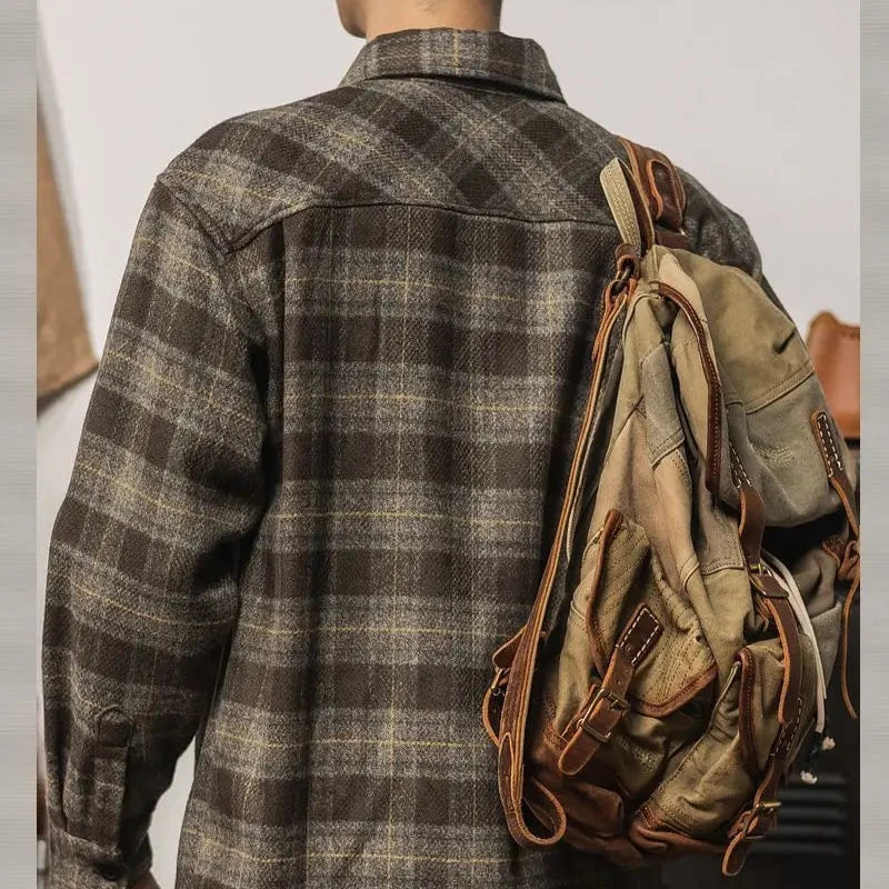 A person wearing a relaxed fit brown plaid flannel shirt, seen from behind, carries a rugged, tan canvas backpack with leather straps over one shoulder.